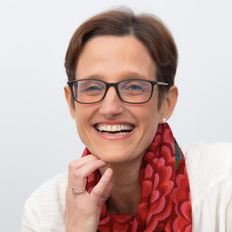 portrait d'une femme tout sourire, cheveux courts, lunettes, écharpe rouge à motif, large bracelet